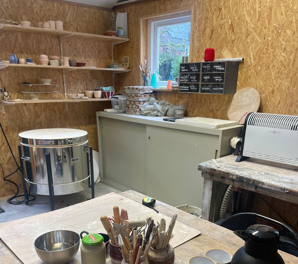 Binnenkant van Atelier Aardwerk met de oven, planken vol handgemaakte keramiek en de werktafel
